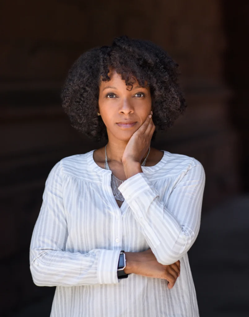 Woman with curly hair in a striped shirt stands thoughtfully, resting her hand on her face, reflecting during the Martin Luther King Jr. Celebration at Valpo featuring Imani Perry.