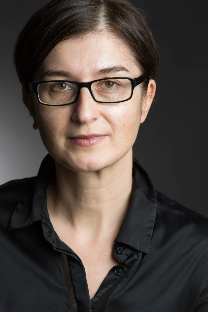 Woman with brown hair, glasses, and black shirt smiles slightly against a dark, neutral background, reflecting her commitment to interdisciplinary development as part of the Baepler Professorship.