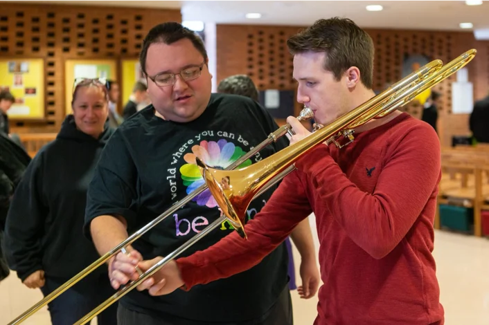 A man helps another man play a trombone indoors, with people watching in the background—part of Valparaiso University’s Sensory Friendly Music Therapy Program.