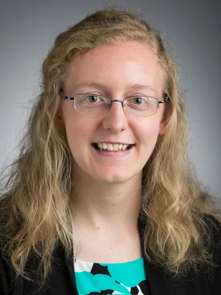 Emily Prough, Employee of the Month in March 2020, smiles at the camera against a gray background, her wavy blonde hair and glasses complementing her stylish black blazer.
