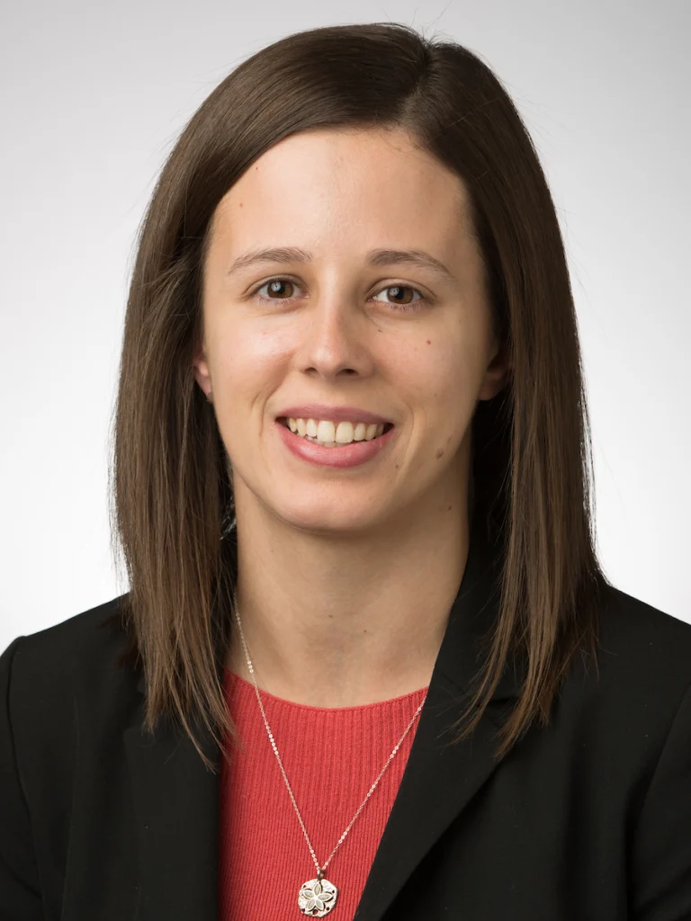Woman with straight brown hair wearing a black blazer and red top, smiling at the camera; her eyes reflect curiosity and passion for learning.