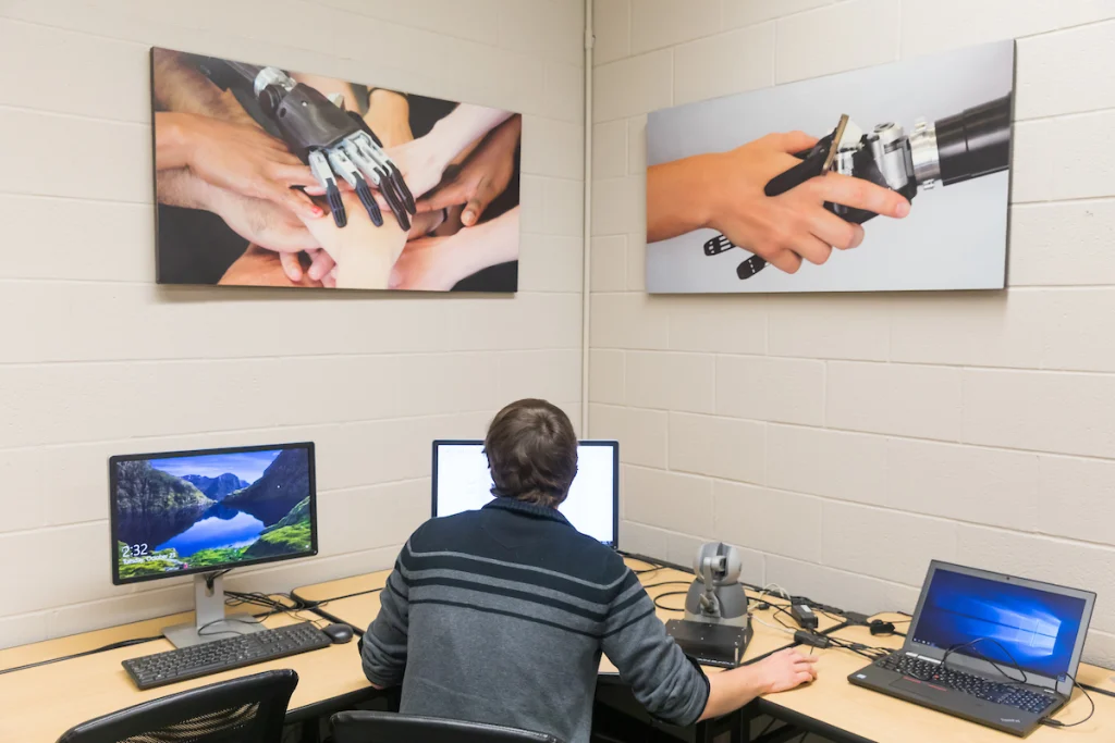 Person at a desk with computers, beneath photos of robotic and human hands collaborating—a nod to mechatronics and the future of engineering automation.