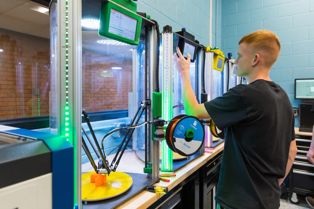 A person operates a 3D printer in an Engineering PLUS lab, adjusting settings on a touchscreen next to filament spools.