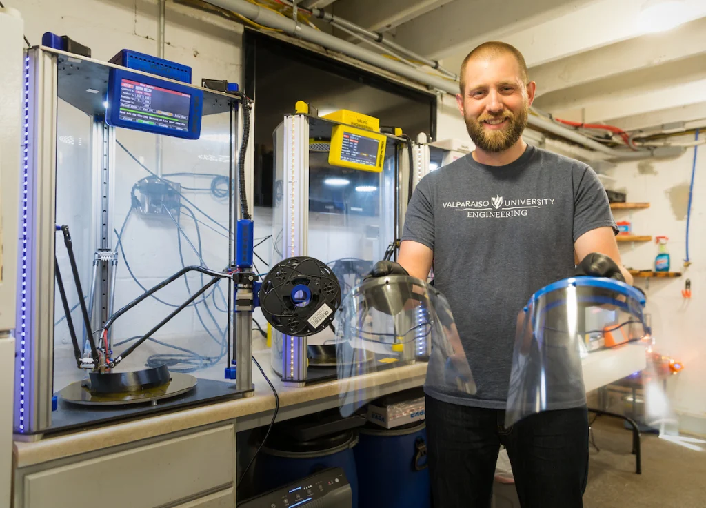 A man holds two face shields by 3D printers in a workshop, smiling at the camera—showcasing the community spirit of Valpo professors.