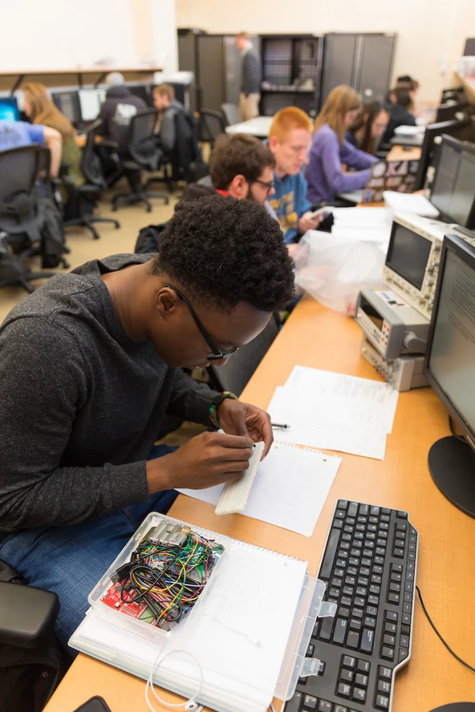 A person assembles electronics at a desk with wires, papers, and a computer in a busy classroom, showcasing hands-on learning experiences supported by the IPC Education Foundation for engineering students.