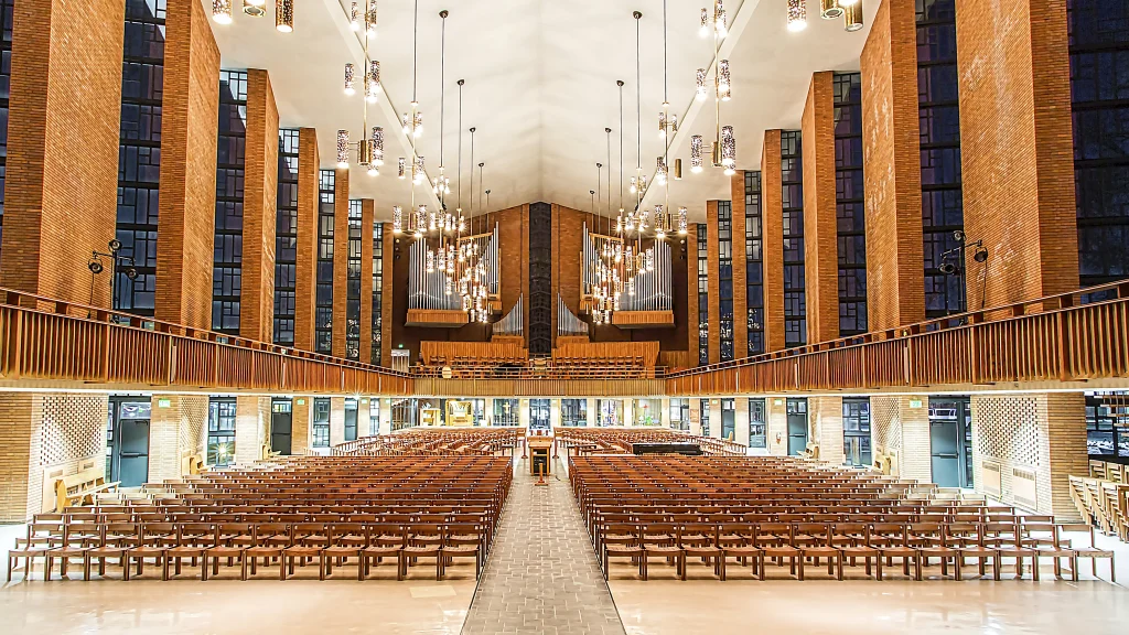 Modern church interior with rows of wooden chairs, tall windows, and hanging lights, viewed from the aisle—an inviting space where Members of the 2020 Class once gathered for special ceremonies.
