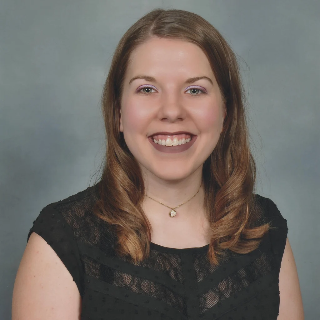 Woman with long brown hair in a black lace top, smiling at the camera against a plain gray background, celebrating with fellow 2020 Class members.