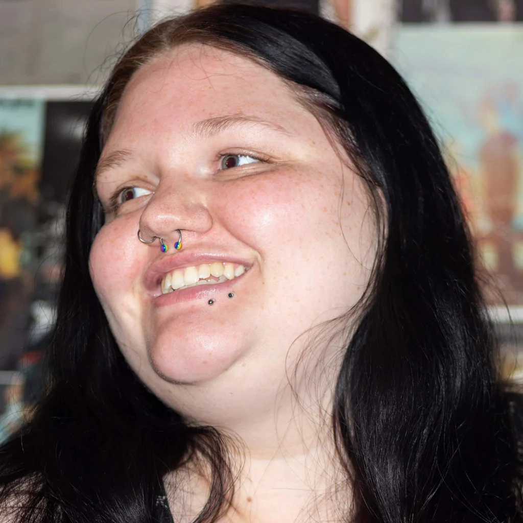 Members of the 2020 Class, this person with long dark hair is smiling, wearing a nose ring and lip piercings, with a blurred background.