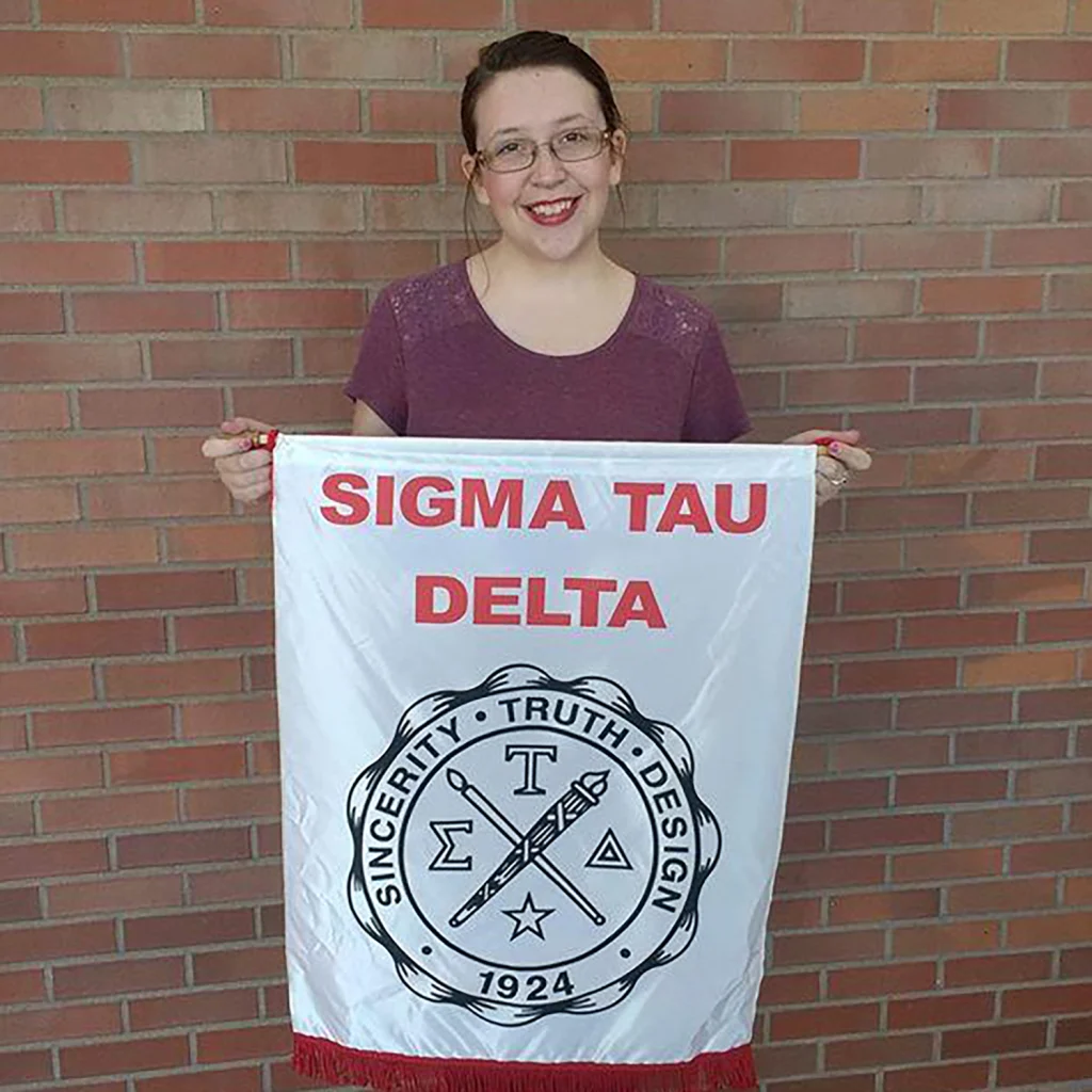 A smiling member of the 2020 Class holds a Sigma Tau Delta banner, standing proudly against a brick wall in the background.