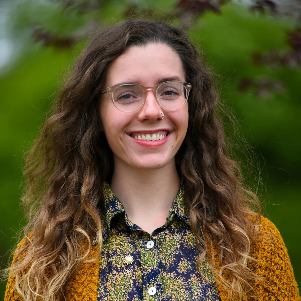Smiling member of the 2020 Class with glasses and long curly hair, wearing a floral shirt and brown sweater, outdoors.