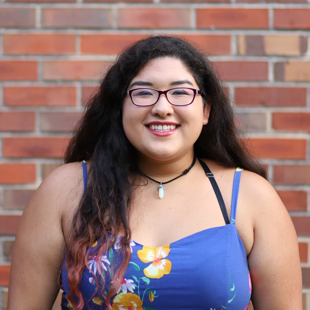 Woman with long dark hair wearing glasses and a blue floral dress, smiling in front of a brick wall—one of the proud Members of the 2020 Class.