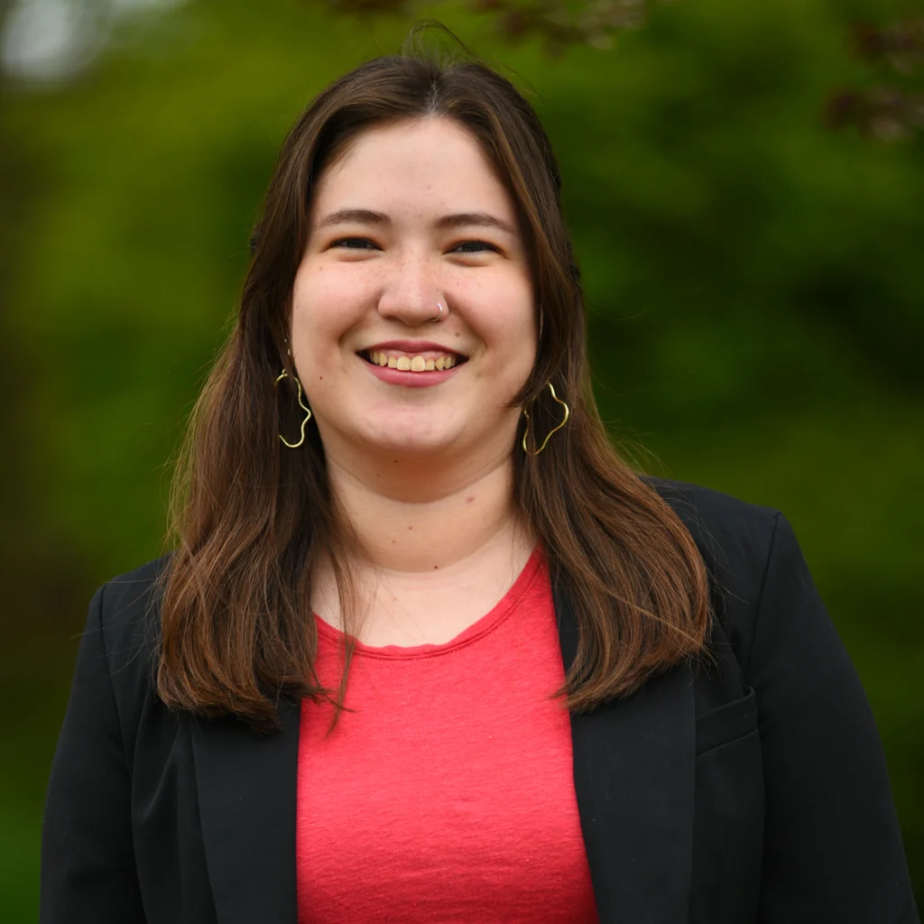 A woman with long brown hair smiles outdoors, wearing a black blazer, red shirt, and abstract earrings—one of the distinguished Members of the 2020 Class.