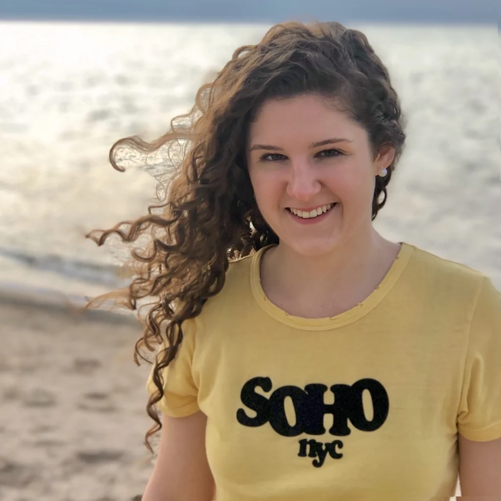 Smiling woman with curly hair in a yellow "SOHO nyc" shirt stands on a beach with water in the background, celebrating with fellow members of the 2020 Class.
