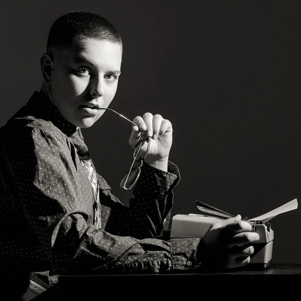 A member of the 2020 Class with short hair holds eyeglasses while sitting at a table with a typewriter, captured in striking black-and-white lighting.