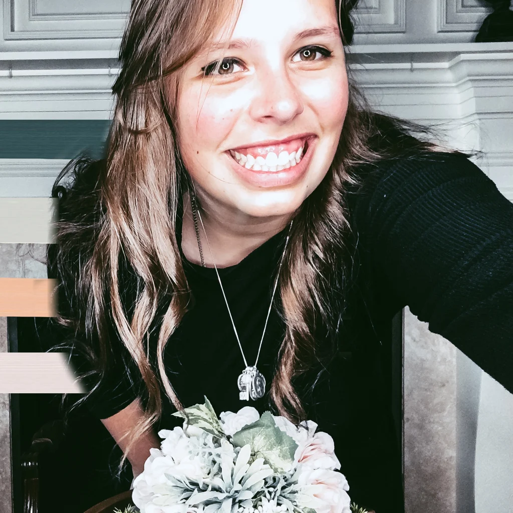Smiling woman with long hair, a member of the 2020 Class, holding white flowers and wearing a black top and a silver necklace indoors.