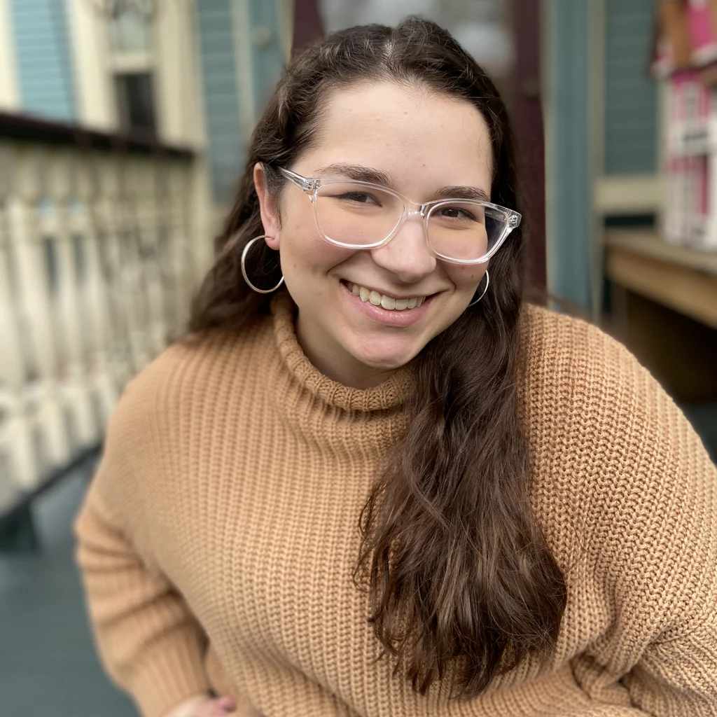 A woman with long brown hair and glasses, a proud member of the 2020 Class, smiles in a tan sweater as she sits on a porch.