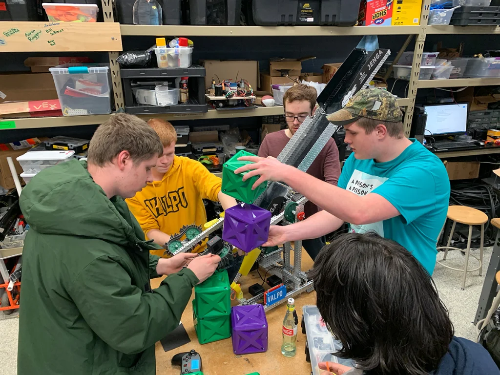 Four students from the Valparaiso University Robotics Team collaborate on a project, handling colorful cubes and mechanical parts on a bench as they prepare for the World Championship.