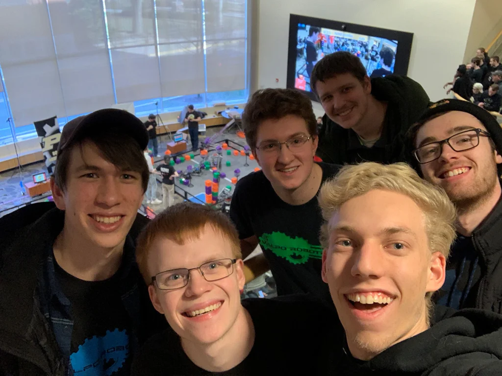 Six smiling young men from the Valparaiso University Robotics Team take a group selfie at a robotics World Championship inside a large, bright room.