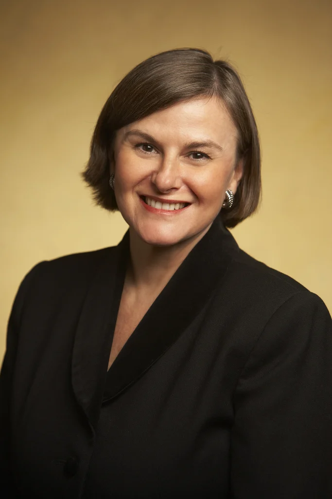 Woman with short brown hair in a black blazer, smiling in front of a plain beige background. She is the Interim President of Valparaiso University.