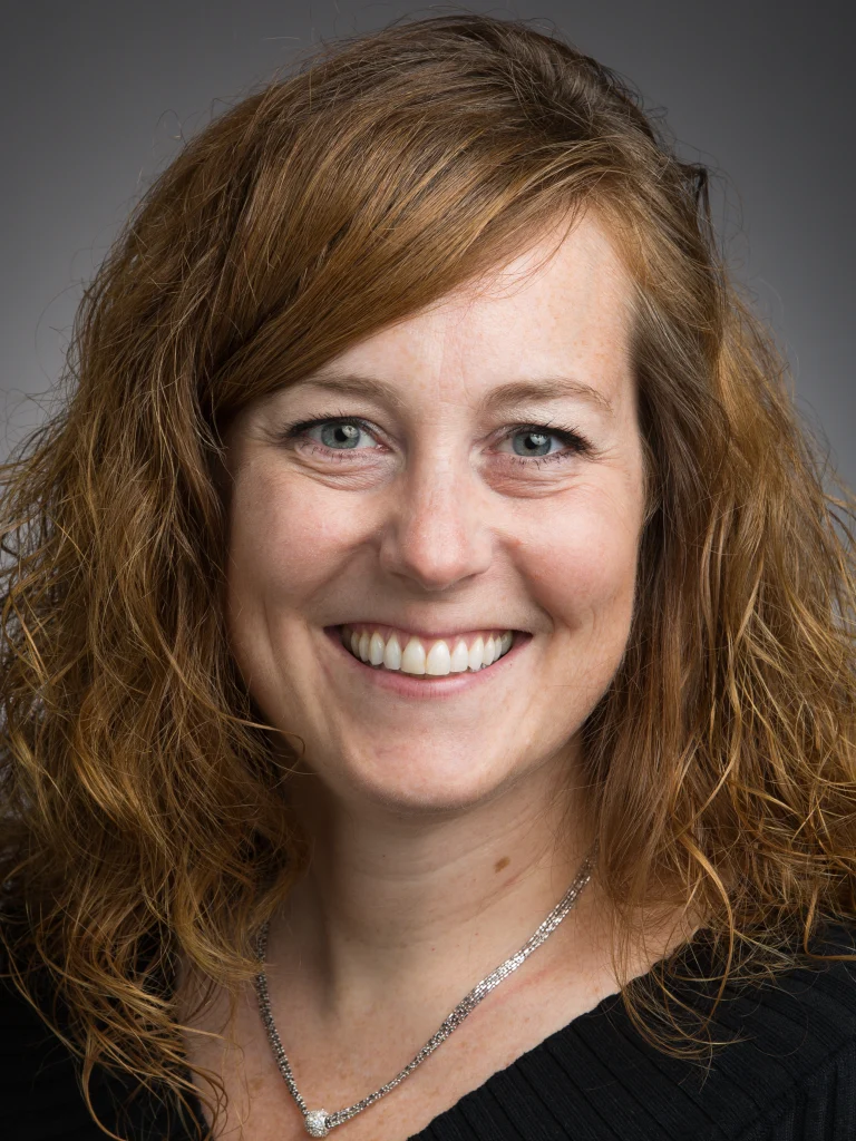 Smiling woman with wavy, light brown hair wearing a black top and silver necklace, pictured against a gray background—an approachable member of the Incident Command Team.