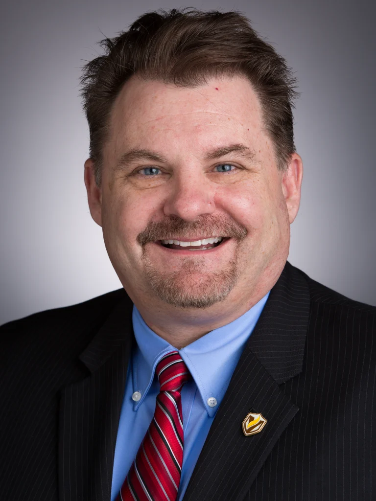 Smiling man in a suit, blue shirt, and red striped tie, with short brown hair and goatee, appearing ready to lead as part of an incident command team.