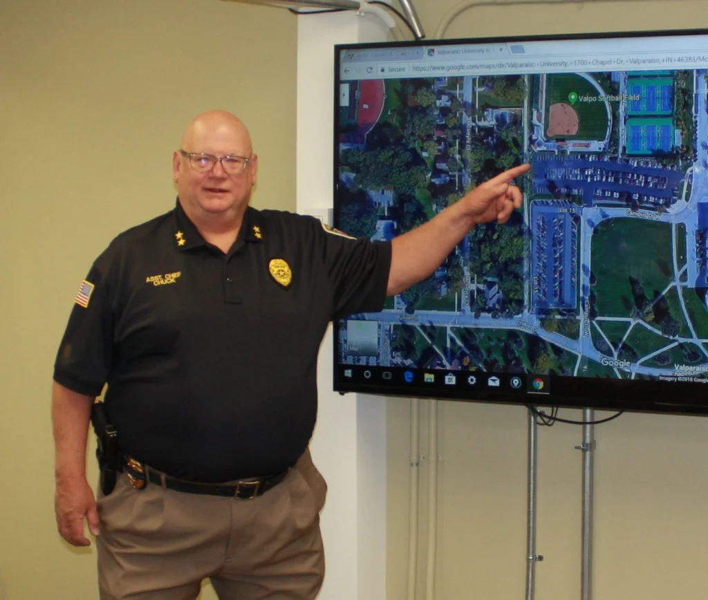 A man in a police uniform points at a map displayed on a large screen, briefing the Incident Command Team in an indoor setting.