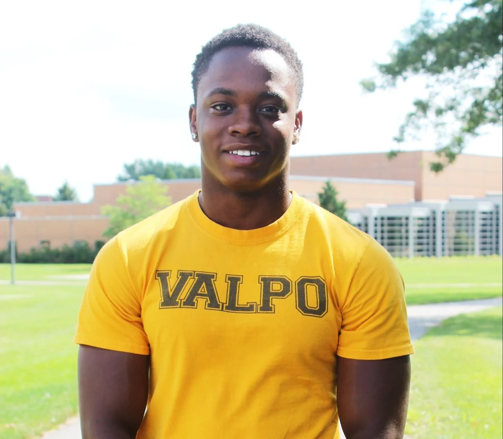 A smiling person wearing a yellow "VALPO" shirt stands outside on a sunny day, embodying advocacy with confidence, with a building in the background.