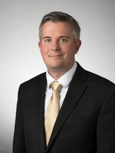 A man in a black suit and gold tie, named SEAC Employee of the Month in July 2020, poses for a professional portrait against a plain background.