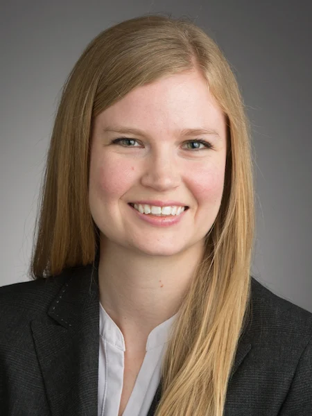 Young woman with long blonde hair, smiling, wearing a black blazer and white shirt, showcasing the poise of an Economics Professor recognized for Excellence in Teaching, against a gray background.