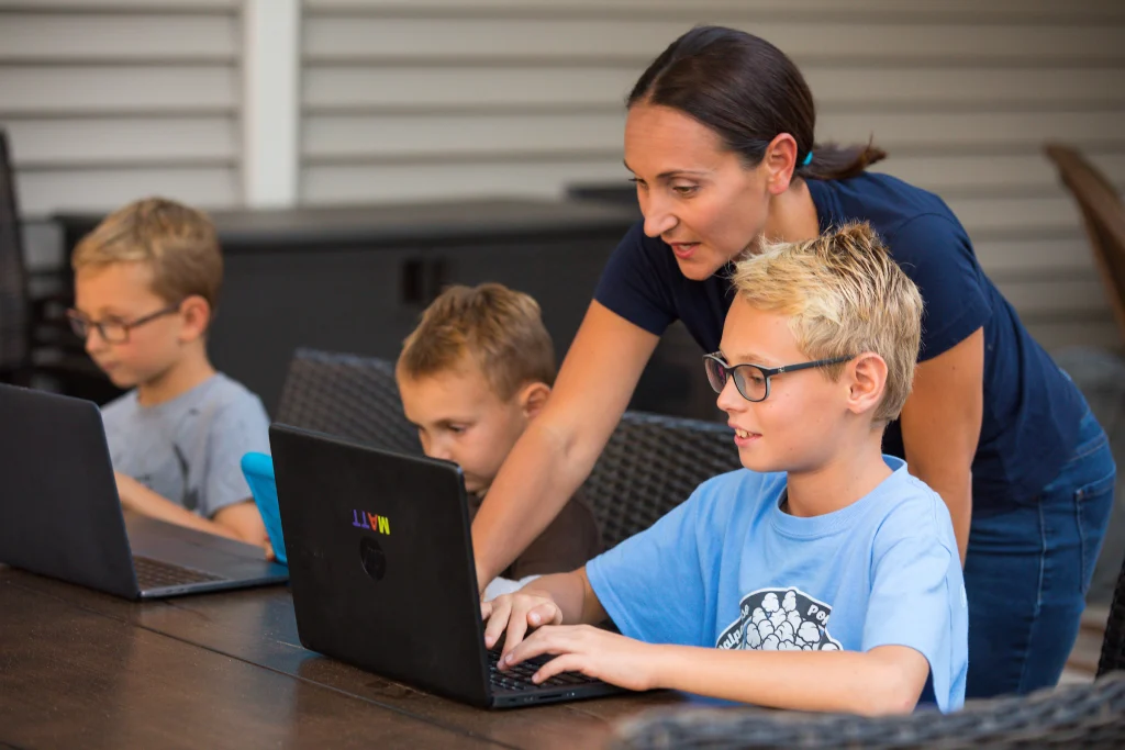 A woman helps three young boys working on laptops at an outdoor table, illustrating how parents cope with remote learning challenges during the pandemic.