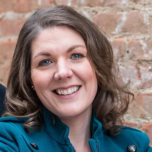 A woman with wavy brown hair smiles, wearing a teal coat, standing in front of a brick wall—proudly posing as SEAC’s Employee of the Month.