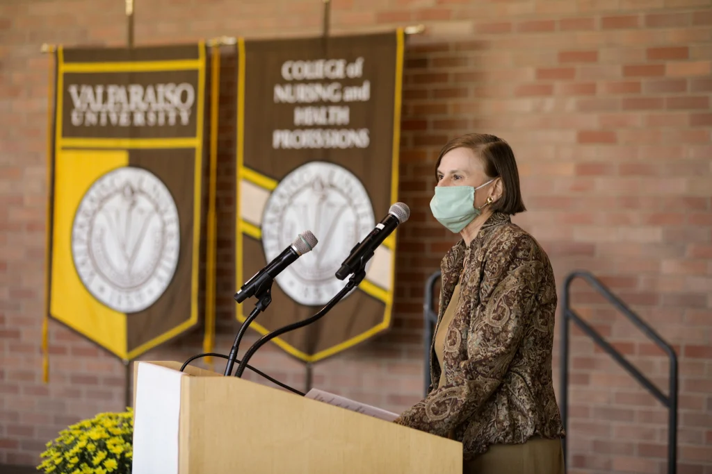 A woman in a mask speaks at a podium with Valpo university and nursing college banners behind her, addressing future Physician Assistant graduates.