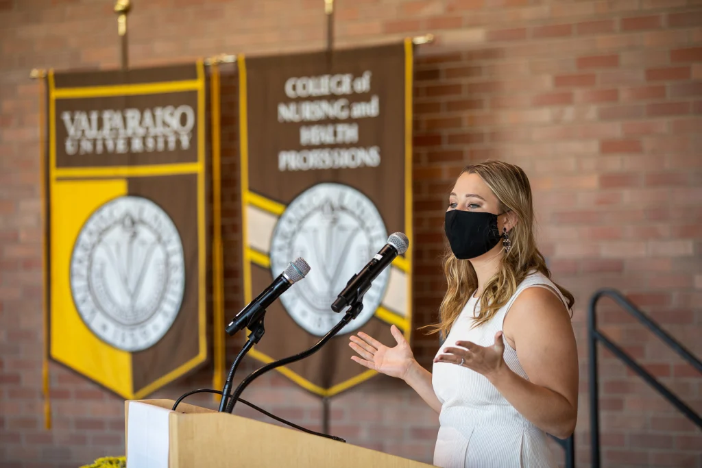 A woman in a mask speaks at a podium with Valpo nursing college banners in the background, addressing graduates and future physician assistant professionals.