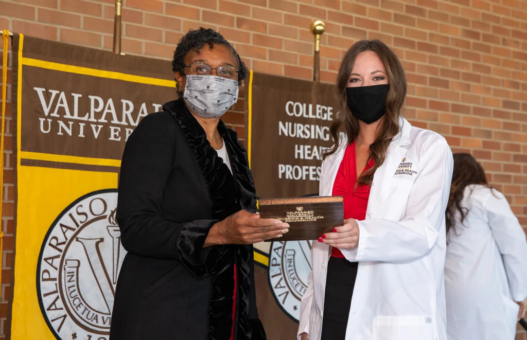 Two women in masks hold a plaque at a Valpo nursing event, standing in front of banners and celebrating the achievements of graduates.