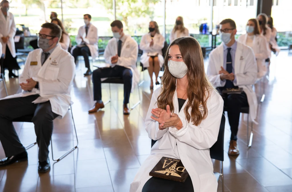 Valpo Physician Assistant graduates in white coats, wearing masks, sit spaced apart and clap at an indoor ceremony with natural light.