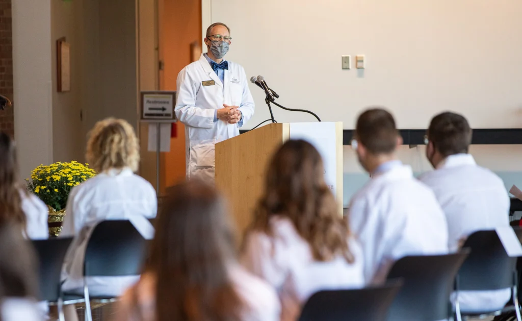A Physician Assistant addresses fellow graduates at a podium, with the Valpo audience also dressed in white coats.