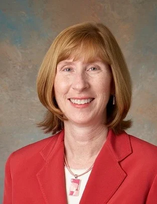 Smiling woman with straight red hair, wearing a red blazer and pendant necklace, stands against a neutral background—Theresa Kessler of Valparaiso University is honored by the American Academy of Nursing.