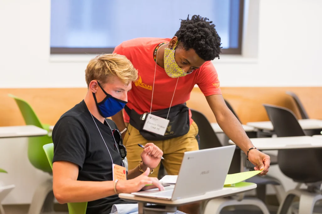 Two students wearing masks work together in a classroom, demonstrating persistence as one helps the other at a laptop on the path to success.