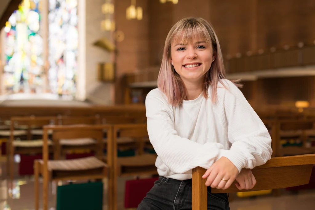 Smiling woman in a white sweater sits on a chair in a sunlit room with stained glass windows, radiating the warmth of faith and community.