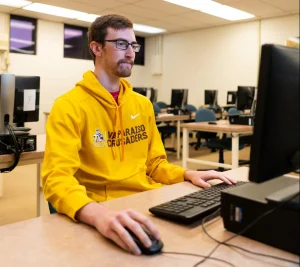 Man in a yellow Valparaiso Crusaders hoodie using a desktop computer in a classroom with empty seats, showcasing the tools for success every aspiring computer scientist needs.