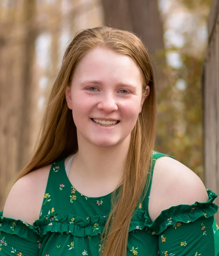 Smiling young woman with long reddish hair in a green floral top, outdoors with trees in the background, proudly displaying her Girl Scouts highest achievement.