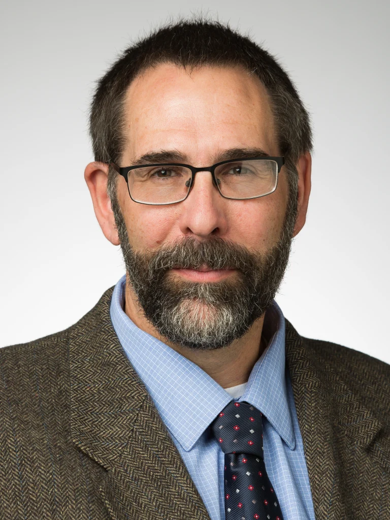Man with glasses, beard, and mustache wearing a brown suit, blue shirt, and patterned tie, Professor Grossman brings professional experience to the classroom; pictured here against a plain background.