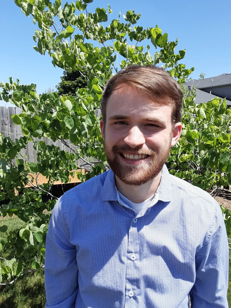 A man with a beard smiles outdoors, standing in front of a leafy green tree on a sunny day, embodying the spirit of Valpo Students and scholars.