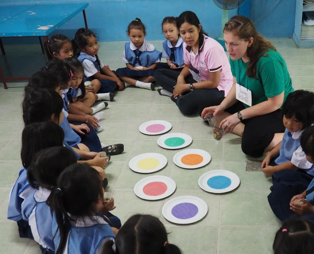 A group of young children sit in a circle with two teachers and colorful paper circles on the floor, exploring their passions and learning about cultures from around the globe.