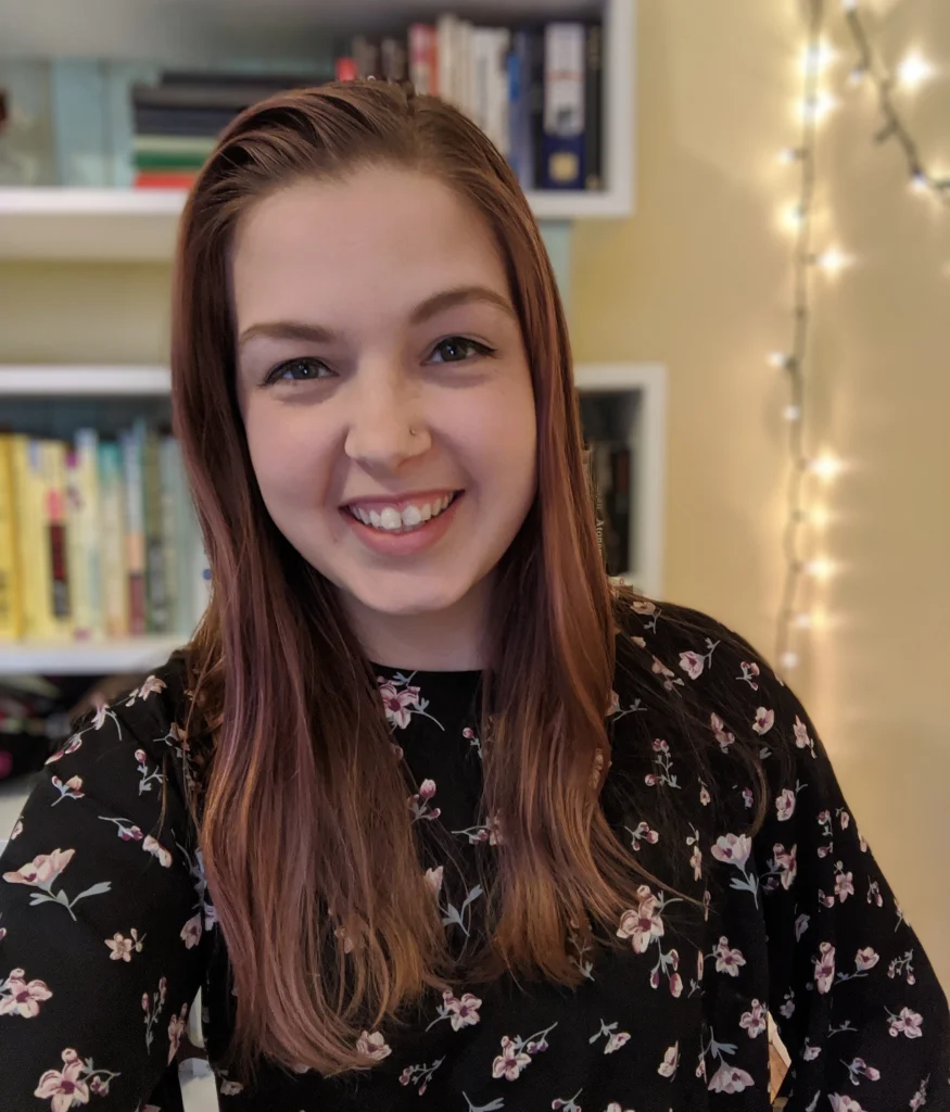 Smiling woman with long brown hair in a floral top, standing indoors by bookshelves and string lights, radiating Creative Connections.