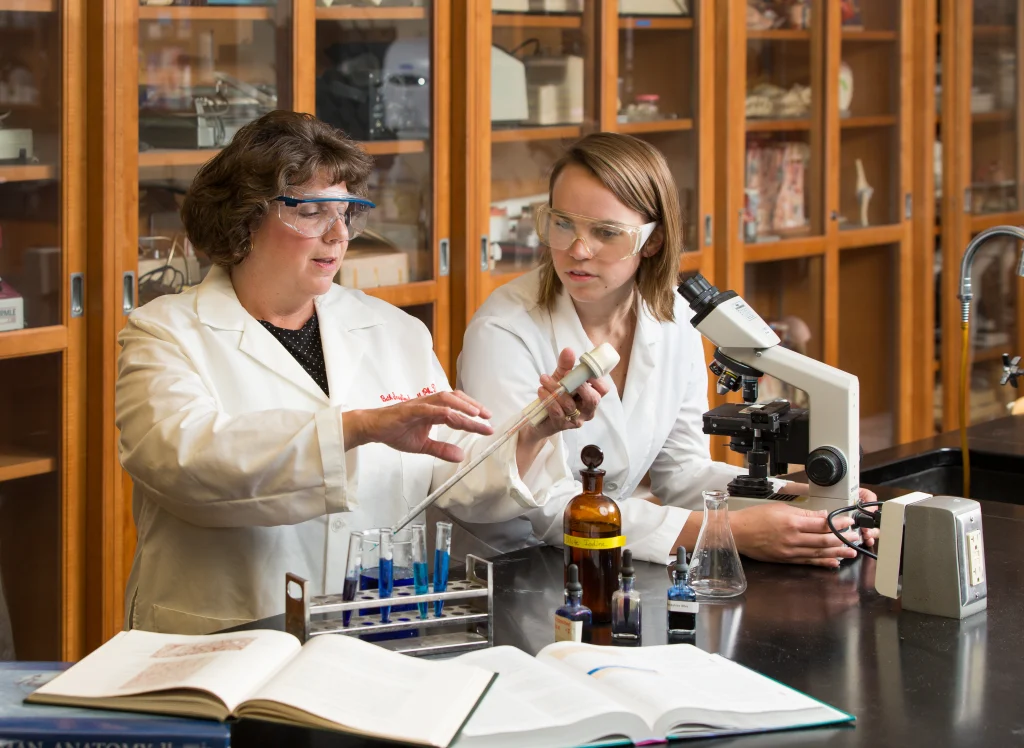 Two women in lab coats offer a helping hand to each other as they work with test tubes and a microscope in a science laboratory.