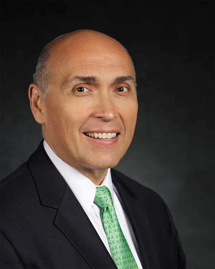 Smiling bald man in a suit, white shirt, and green tie against a dark background, portraying the new president of Valparaiso University.