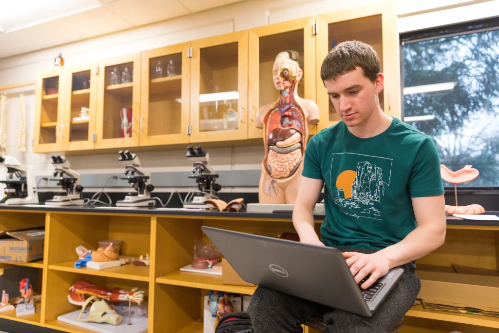 A student uses a laptop in a science classroom with microscopes and anatomical models in the background, preparing for a future among public health graduates who make an impact during challenges like the pandemic.