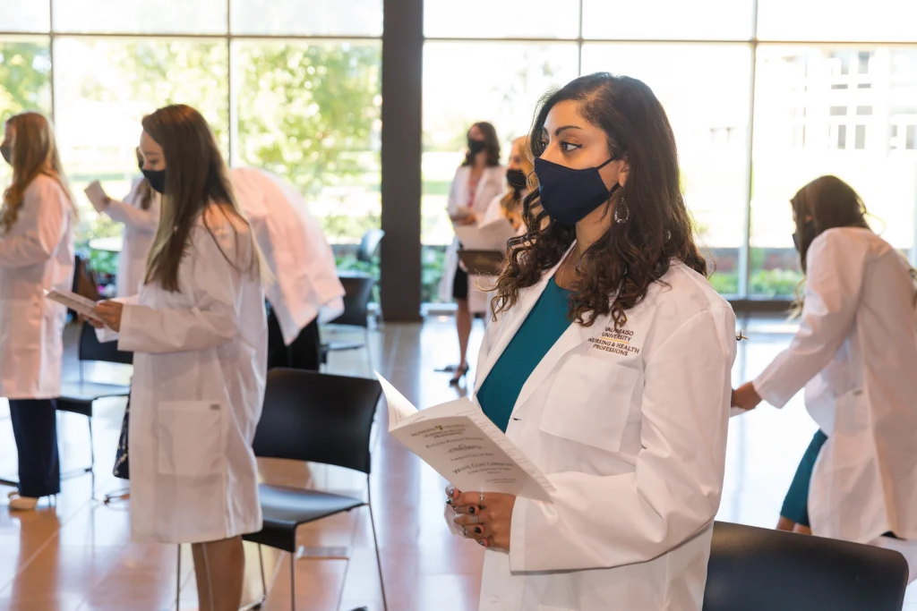 A Valpo woman in a lab coat and mask holds a program at an indoor ceremony, celebrating the First Class of MSPA among others in the background.