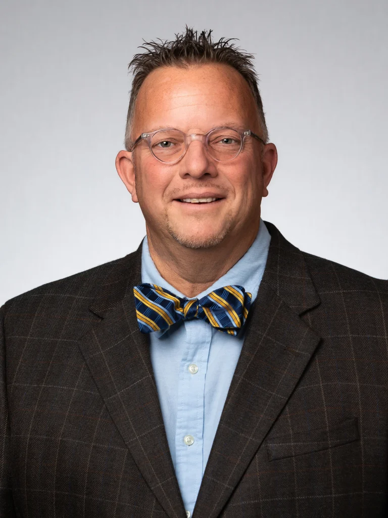 Smiling man with glasses, a plaid jacket, and a blue and yellow striped bow tie stands against a gray background, representing Valpo’s vibrant student life.